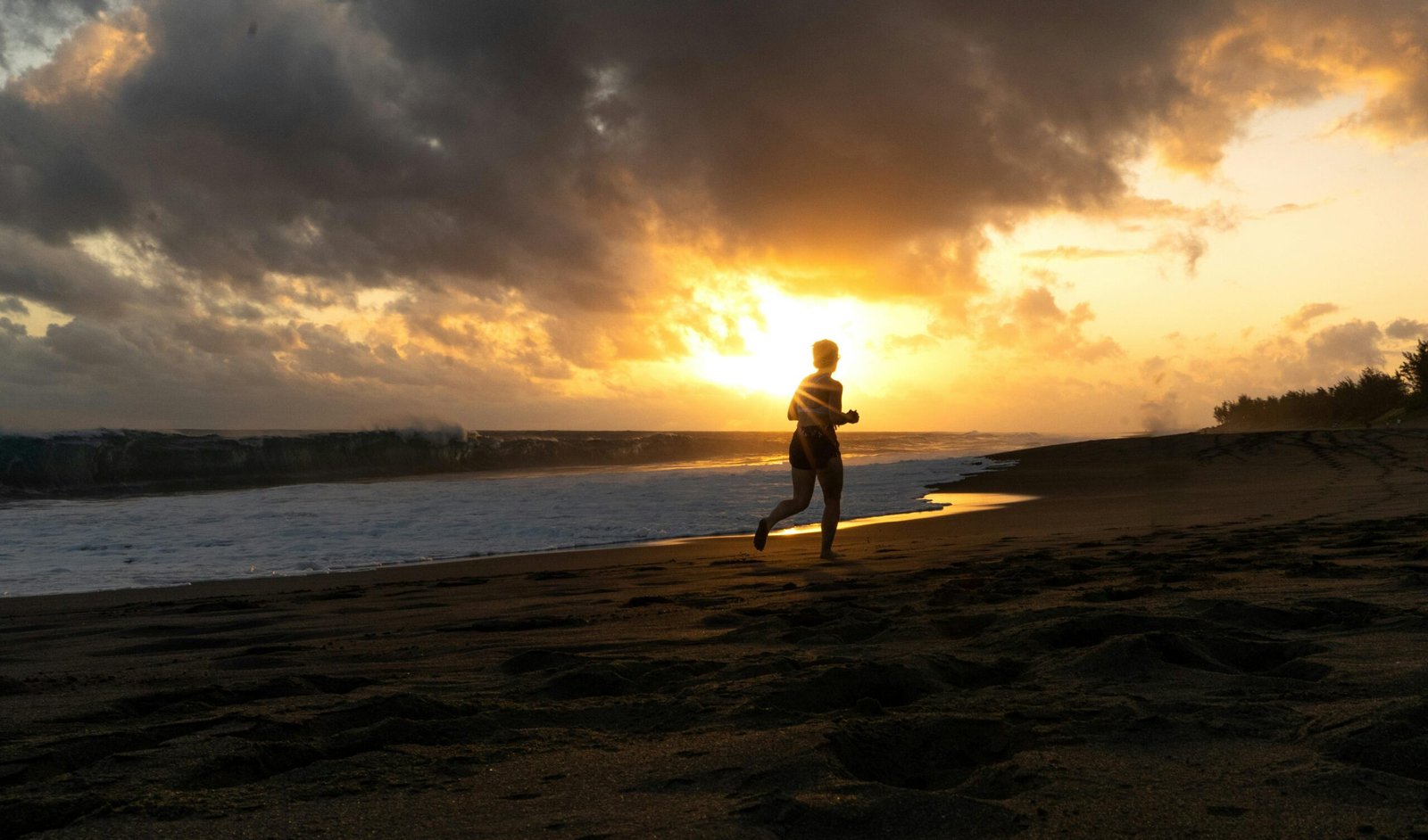 Person jogging on a beach at sunset with ocean waves and glowing sky.