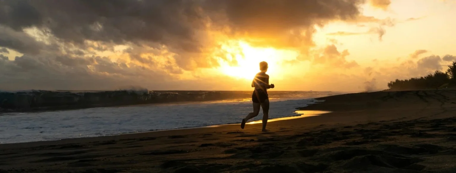 Person jogging on a beach at sunset with ocean waves and glowing sky