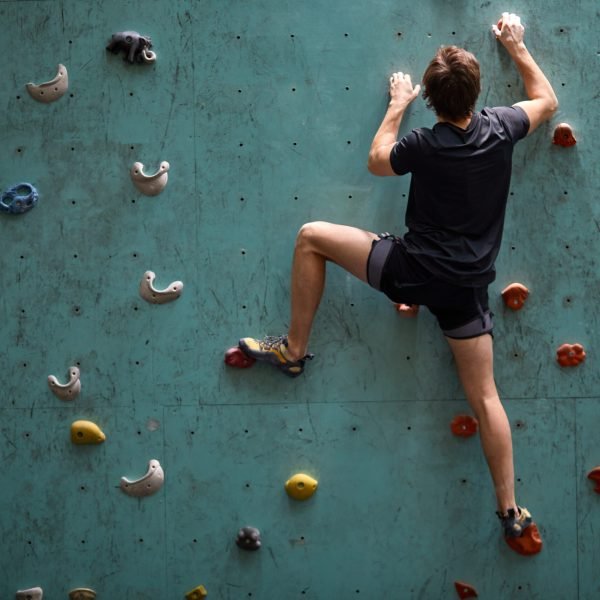 active young caucasian man on rock wall in sport center, rock climbing. Sportive male guy in black sportswear engaged in sport, fitness, lead healthy lifestyle. Rear view