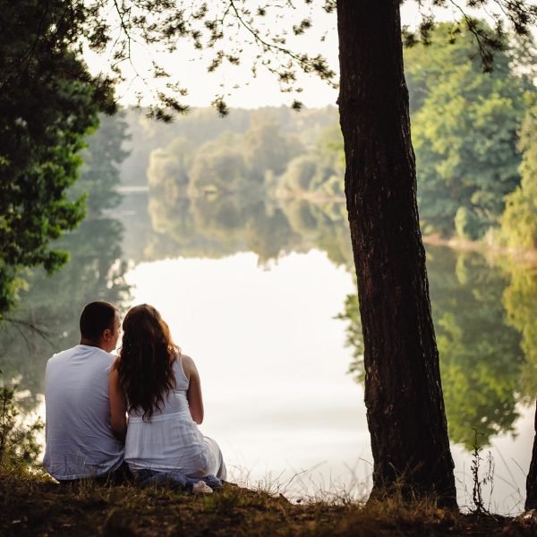 Couple sits and looks at a beautiful sunset near the river. Man and woman look at the river and enjoy nature. Back view of lovely couple man and woman sitting near rivers hugging and relaxing.